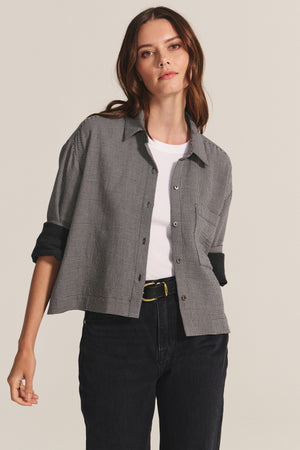 A woman wears the ADELINE SHIRT by Velvet by Graham & Spencer—a black and white houndstooth top—layered over a white tee, with black jeans and a black belt, standing against a plain background.