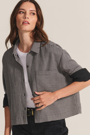 A woman wearing the ADELINE SHIRT by Velvet by Graham & Spencer over a white t-shirt, with black pants and a black belt, stands against a neutral background.