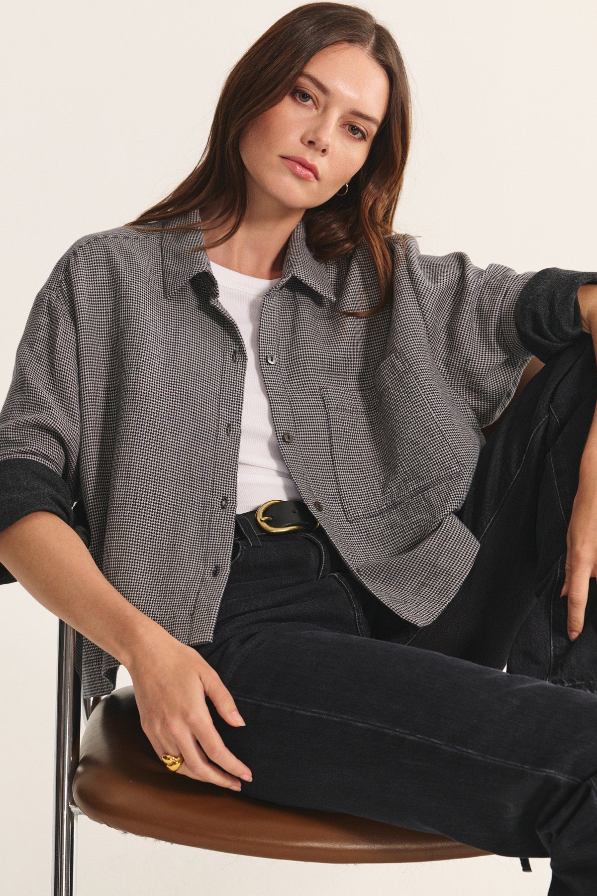 A woman sits on a chair wearing the ADELINE SHIRT by Velvet by Graham & Spencer over a white top with black jeans, one arm resting on her knee and a neutral expression.