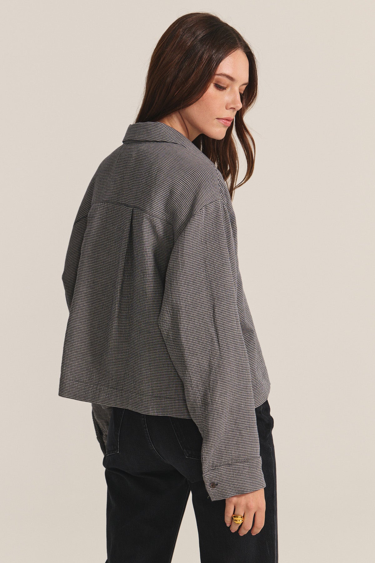 A woman with long brown hair wears the ADELINE SHIRT by Velvet by Graham & Spencer as a loose gray houndstooth jacket with dark pants, standing sideways and looking down against a plain background.