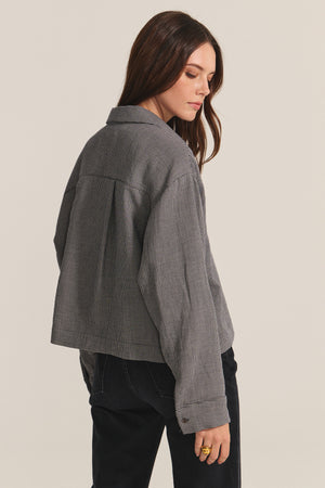 A woman with long brown hair wears the ADELINE SHIRT by Velvet by Graham & Spencer as a loose gray houndstooth jacket with dark pants, standing sideways and looking down against a plain background.