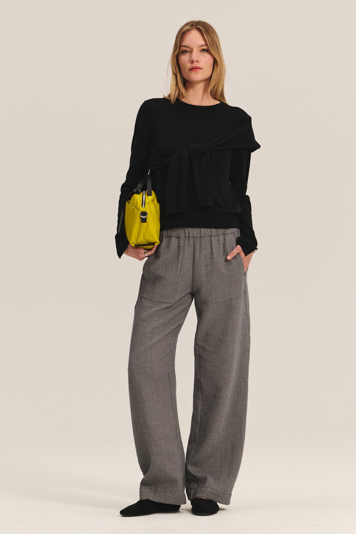 A woman stands against a plain background wearing a black long-sleeve top, Velvet by Graham & Spencer BRAYLEN PANT in cotton gauze with wide-leg silhouette and elastic waistband, black shoes, and holding a small yellow handbag.-40334106394817