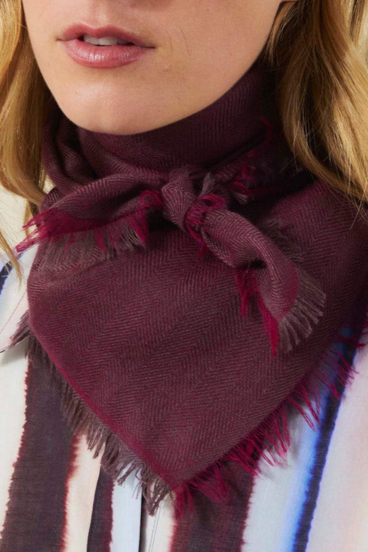 A deep plum-colored SUMMER CASHMERE HERRINGBONE SCARF by Velvet by Graham & Spencer with a herringbone pattern and frayed edges, tied loosely around a person’s neck.