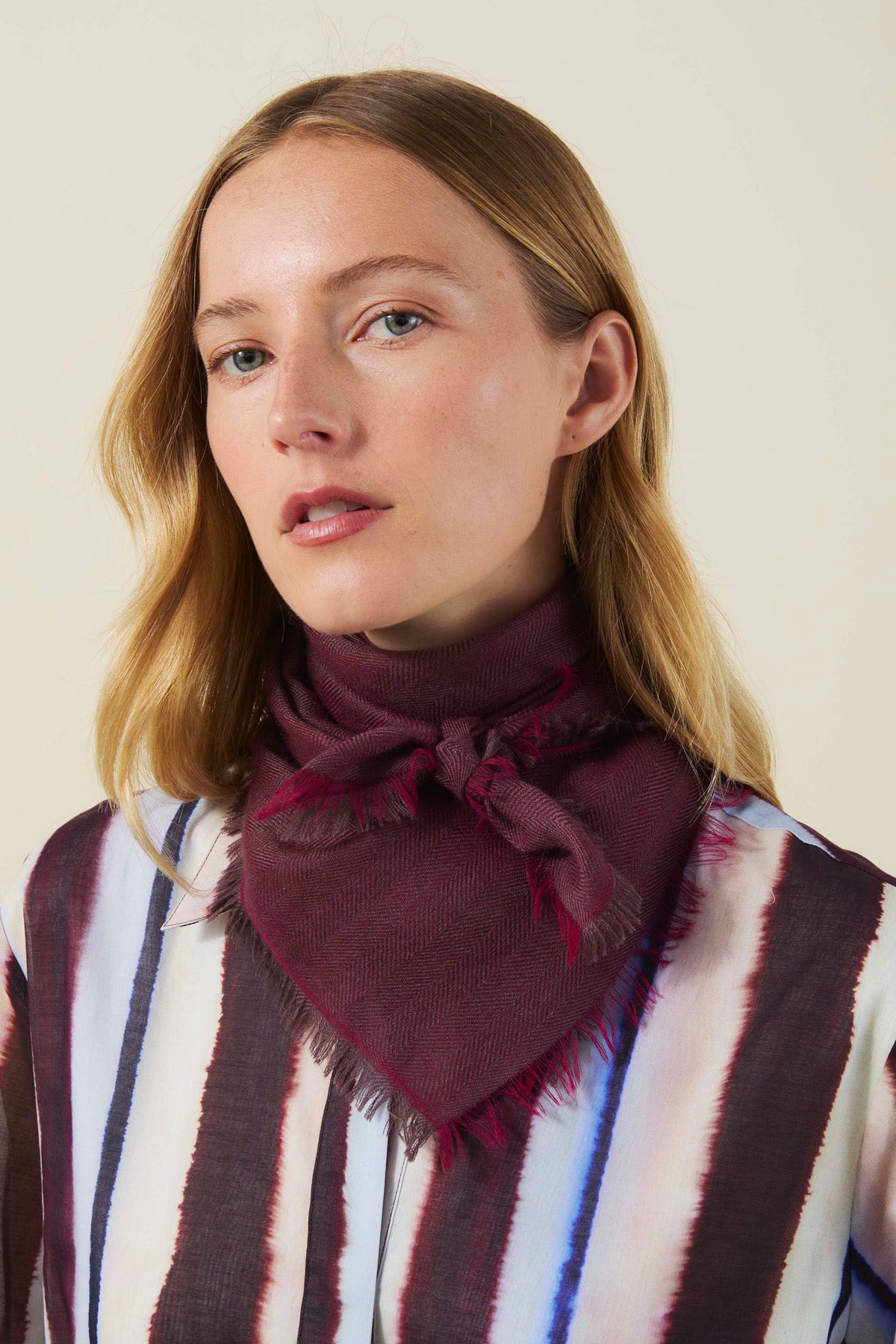 A woman wearing a dark maroon triangular SUMMER CASHMERE HERRINGBONE SCARF by Velvet by Graham & Spencer with a herringbone pattern and frayed edges.