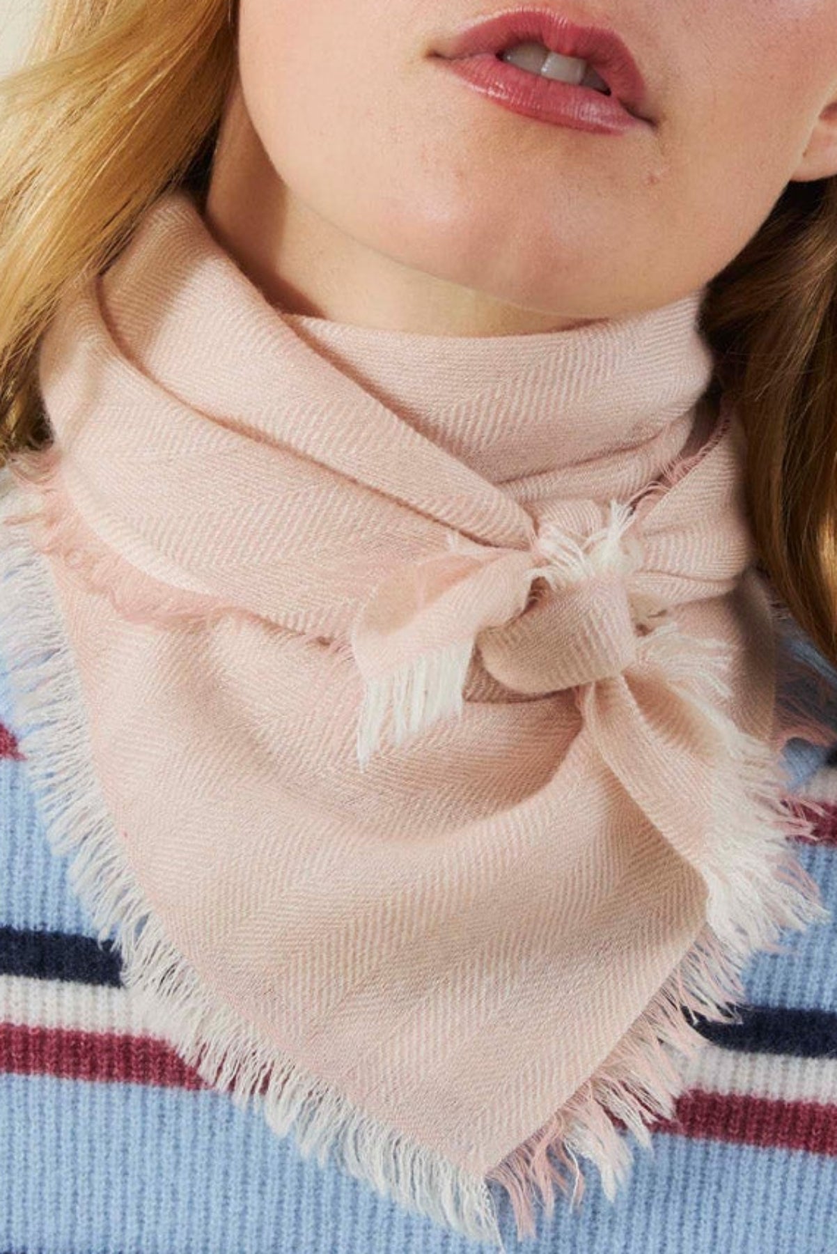 A light pink SUMMER CASHMERE HERRINGBONE SCARF by Velvet by Graham & Spencer, featuring a herringbone pattern and frayed edges, tied loosely around a person’s neck.
