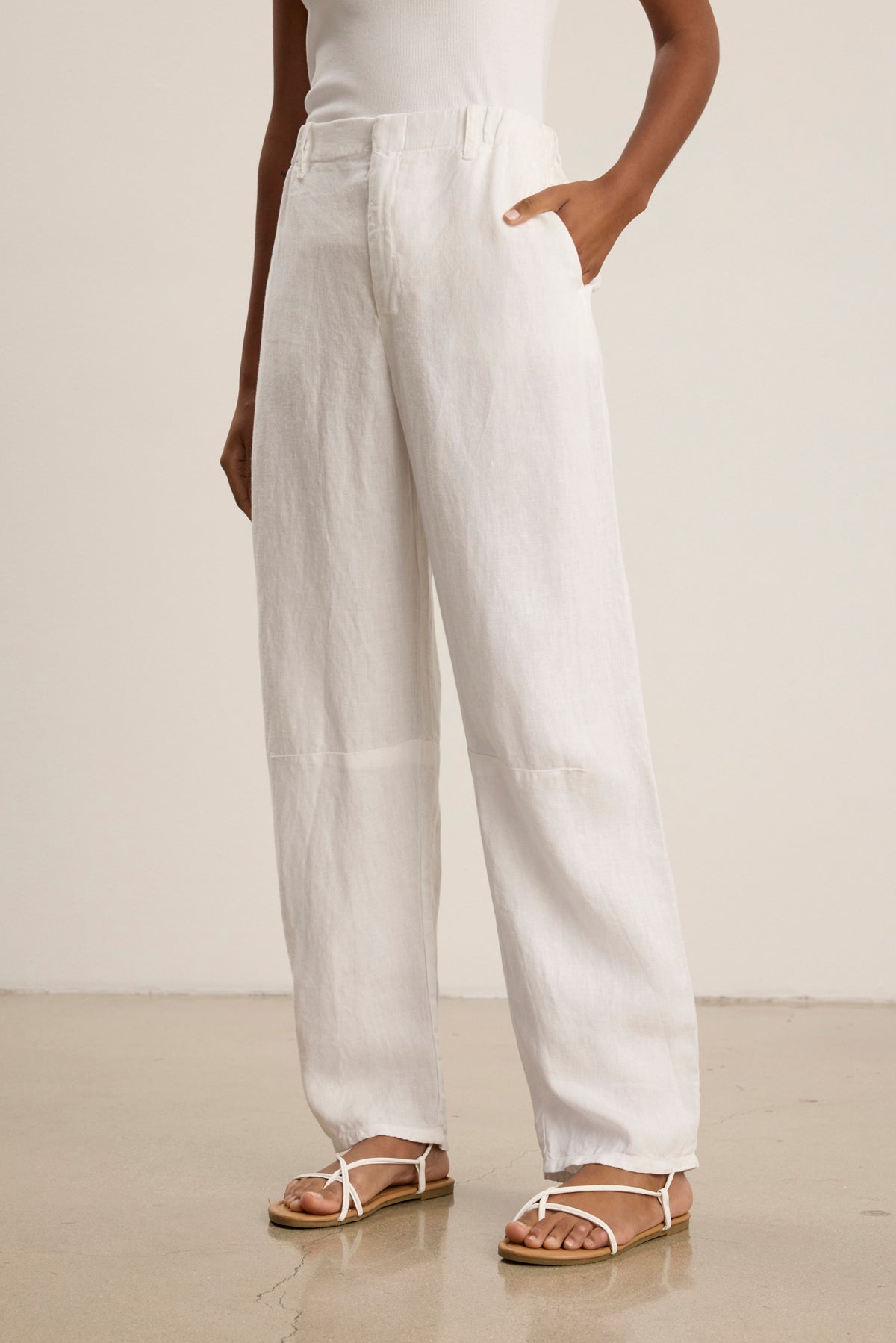 A person stands on a polished floor with one hand in their pocket, wearing Velvet by Graham & Spencer's TAYLOR HEAVY LINEN PANT in white, paired with a white sleeveless top and white sandals.-39111849869505