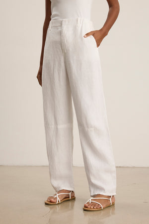 A person stands on a polished floor with one hand in their pocket, wearing Velvet by Graham & Spencer's TAYLOR HEAVY LINEN PANT in white, paired with a white sleeveless top and white sandals.