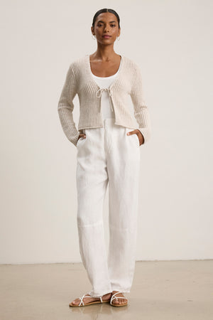 A person stands against a plain background wearing a cream knit cardigan, white tank top, Velvet by Graham & Spencer’s TAYLOR HEAVY LINEN PANT with a tailored fit, and white strappy sandals.