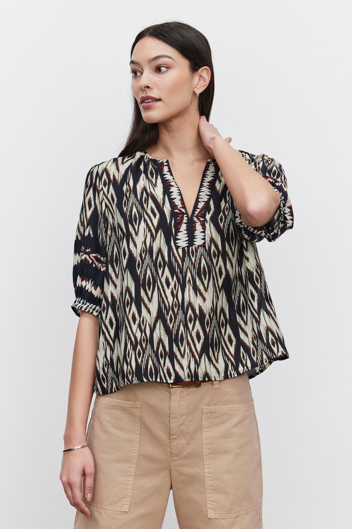   A person with long dark hair wears a BEATRICE TOP featuring an ikat print in luxurious viscose from Velvet by Graham & Spencer, paired with beige pants, standing against a plain white background. 