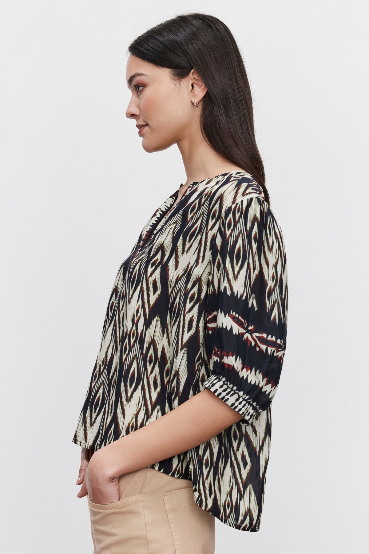   A woman with long dark hair is wearing a BEATRICE TOP by Velvet by Graham & Spencer, crafted from luxurious viscose in an ikat print, paired with beige pants. She stands in a side profile pose against a plain white background. 