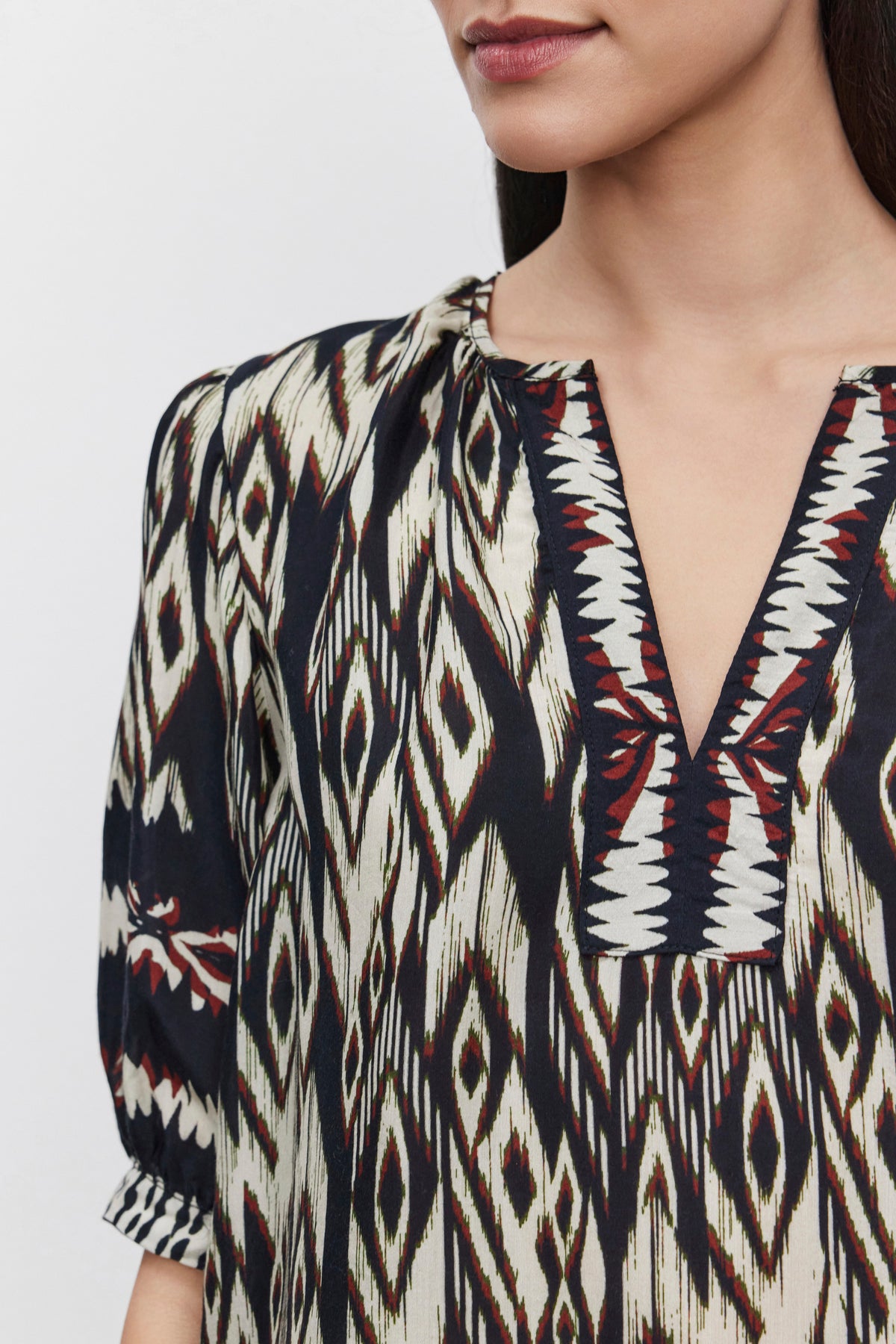   Close-up of a person wearing Velvet by Graham & Spencer's BEATRICE TOP, showcasing an ikat print with a geometric design and a V-neckline. The luxurious viscose fabric enhances their long dark hair, partially visible against a plain white background. 