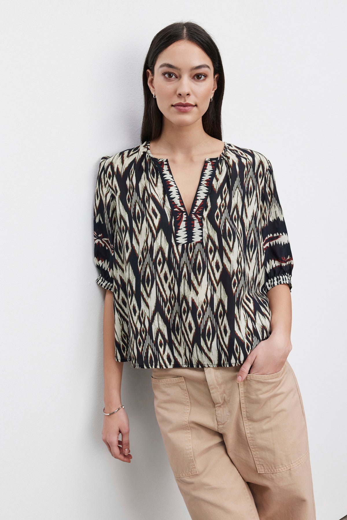   A woman wearing a luxurious Velvet by Graham & Spencer BEATRICE TOP with an ikat print and beige pants stands against a plain white background with one hand in her pocket. 