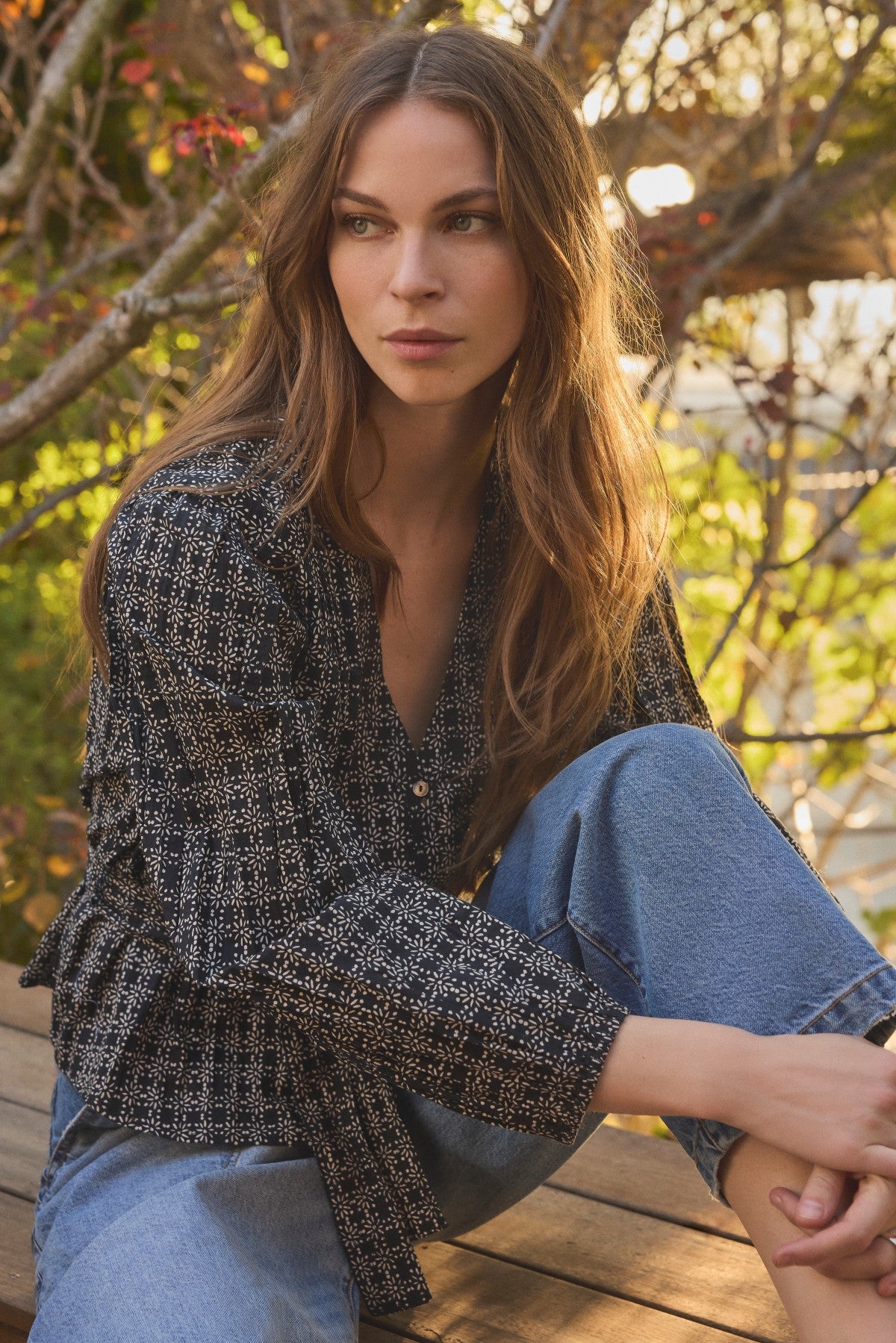A woman with long brown hair sits outdoors on a wooden bench surrounded by trees and sunlight, wearing the IMANI BLOUSE by Velvet by Graham & Spencer paired with blue jeans.