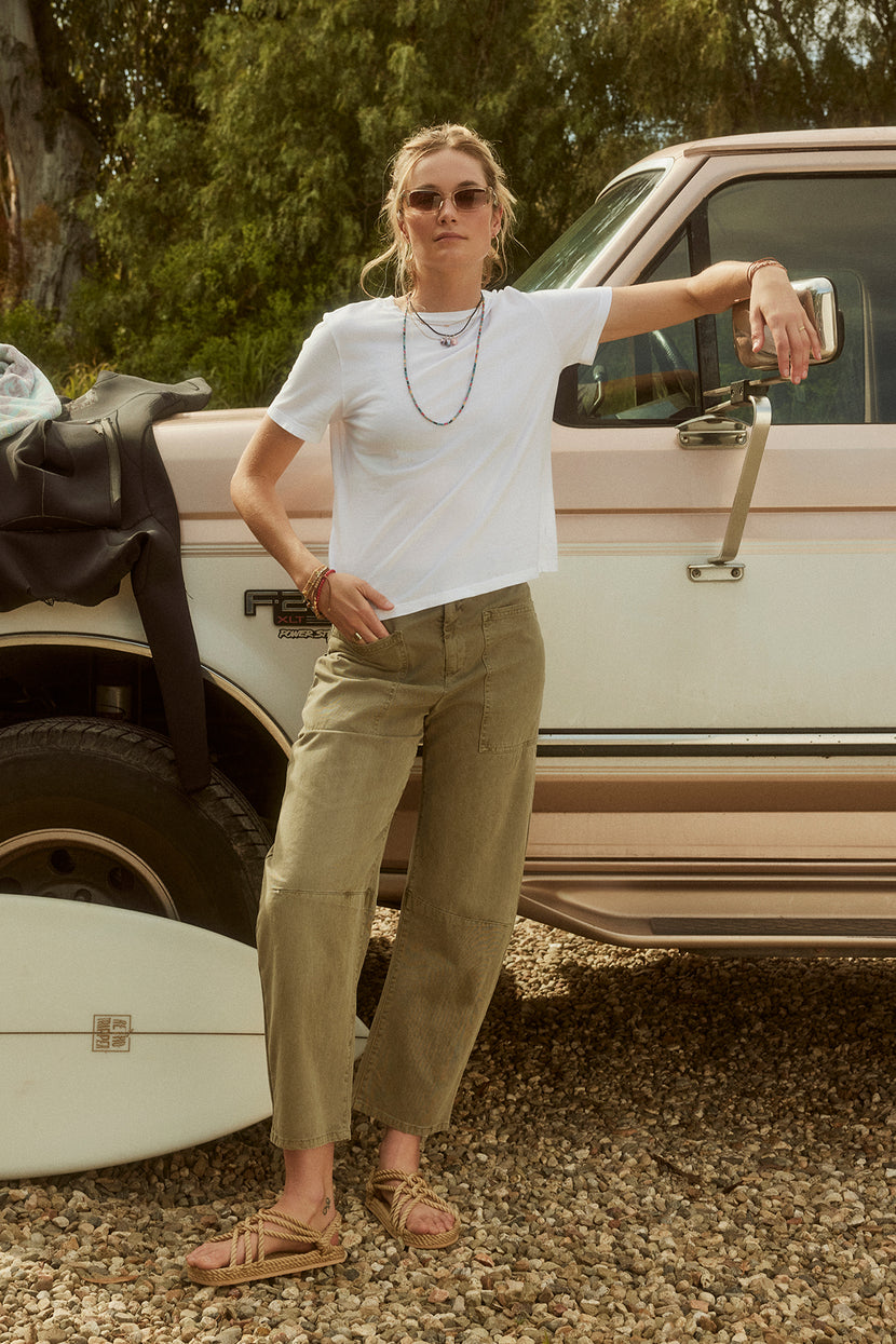 Woman in sunglasses, white t-shirt, and Velvet by Graham & Spencer BRYLIE SANDED TWILL UTILITY PANT leaning on a vintage pickup truck in a gravel area.
