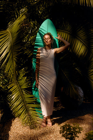 A woman in a white CALLEN DRESS leans against a turquoise surfboard surrounded by tropical plants by Velvet by Graham & Spencer.