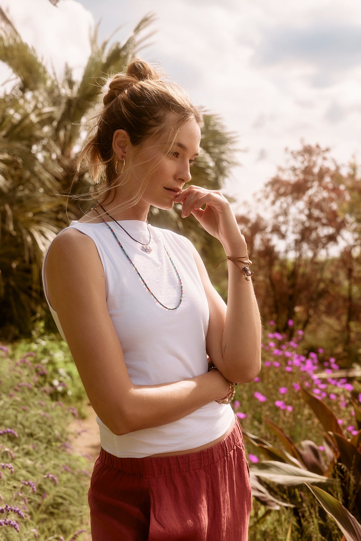   A woman in a Velvet by Graham & Spencer EMILIA TANK TOP and red pants stands pensively in a sunny field with purple flowers, her hand resting on her chin. 