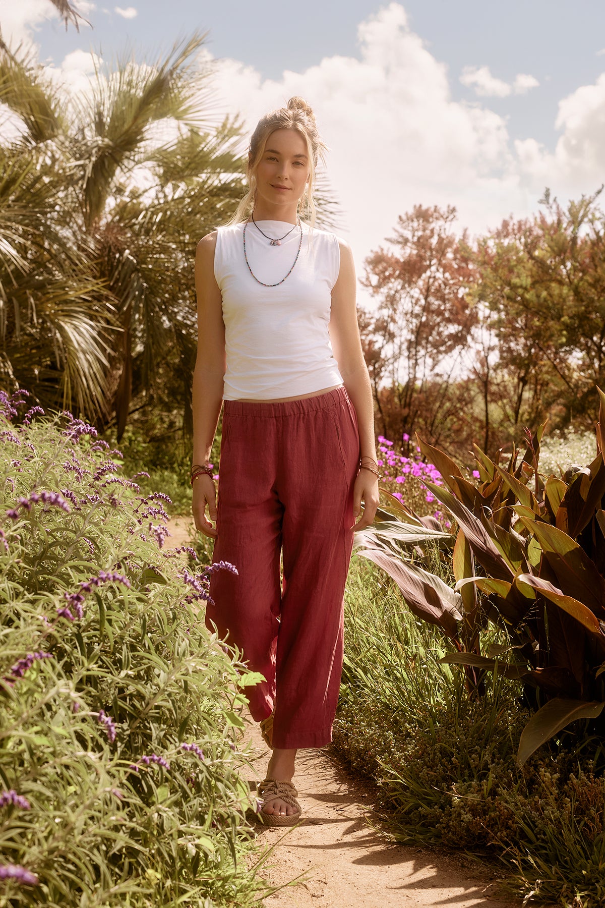   A woman in an EMILIA TANK TOP by Velvet by Graham & Spencer and red pants standing on a garden path surrounded by purple flowers and greenery, under a sunny sky. 