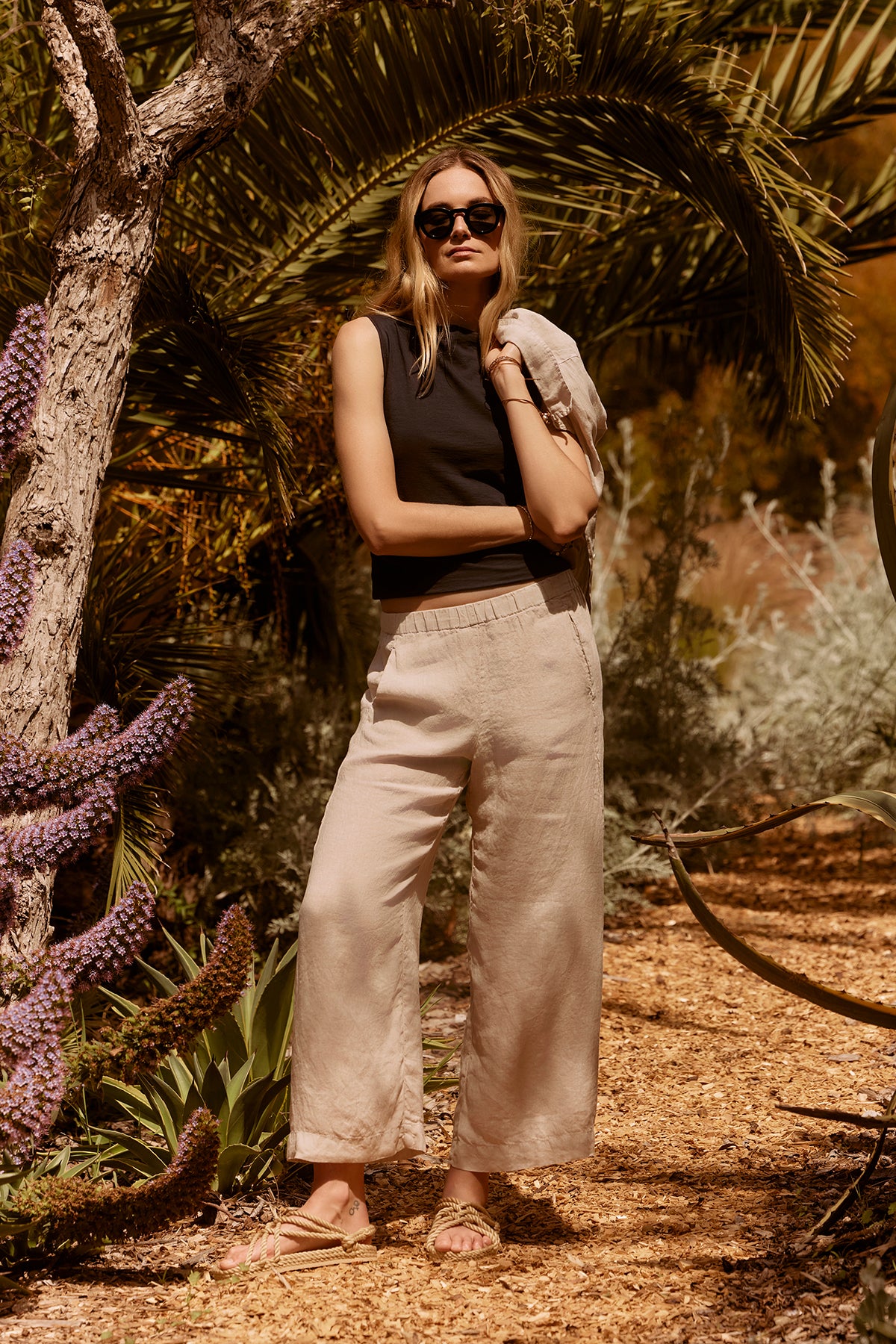 A person wearing sunglasses, a sleeveless black top, and lightweight Velvet by Graham & Spencer LOLA LINEN PANT stands on a dirt pathway surrounded by plants and trees in a sunny outdoor setting.-37000573485249