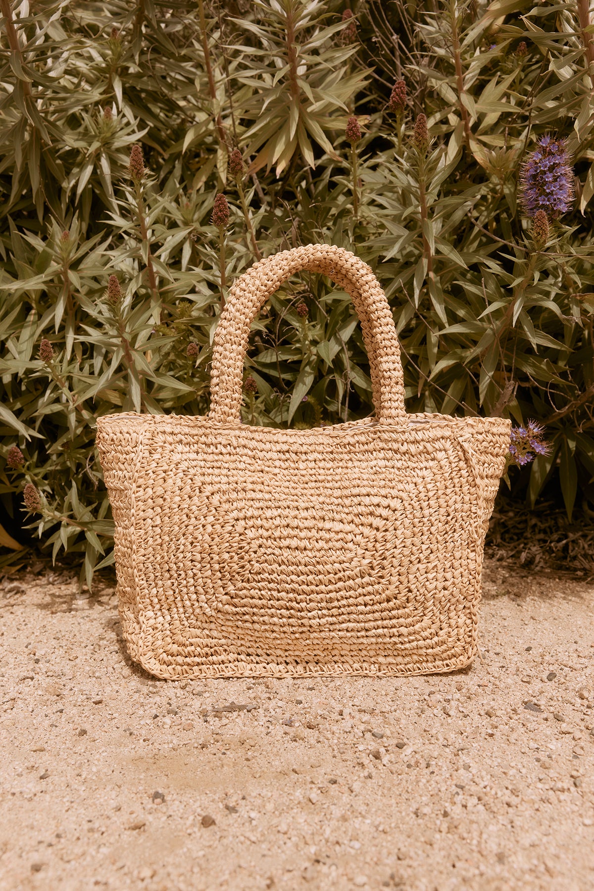   Woven raffia TINA STRAW TOTE BAG by Velvet by Graham & Spencer sits on sandy ground against a backdrop of leafy and flowering plants. 