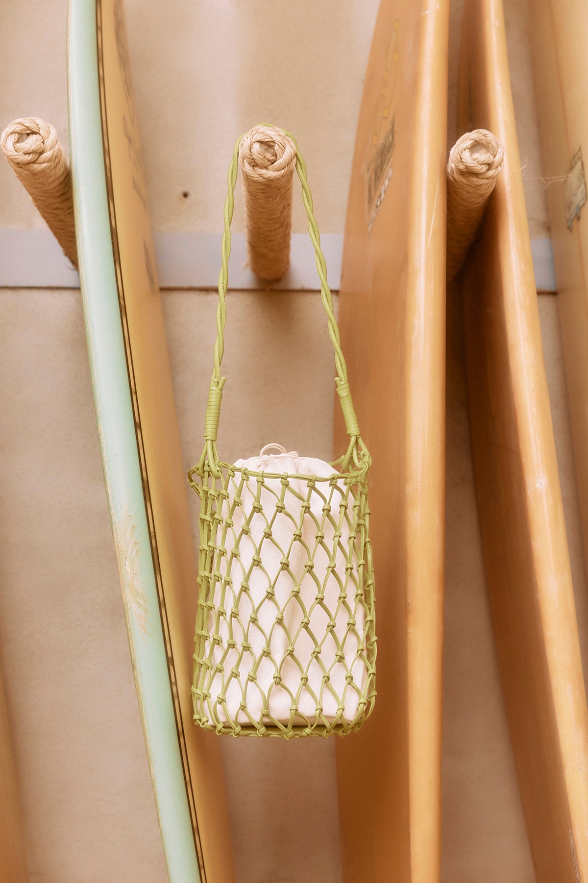 A green Velvet by Graham & Spencer mesh tote bag hanging from a wooden bamboo pole, with other bamboo poles in the background.-37000750858433