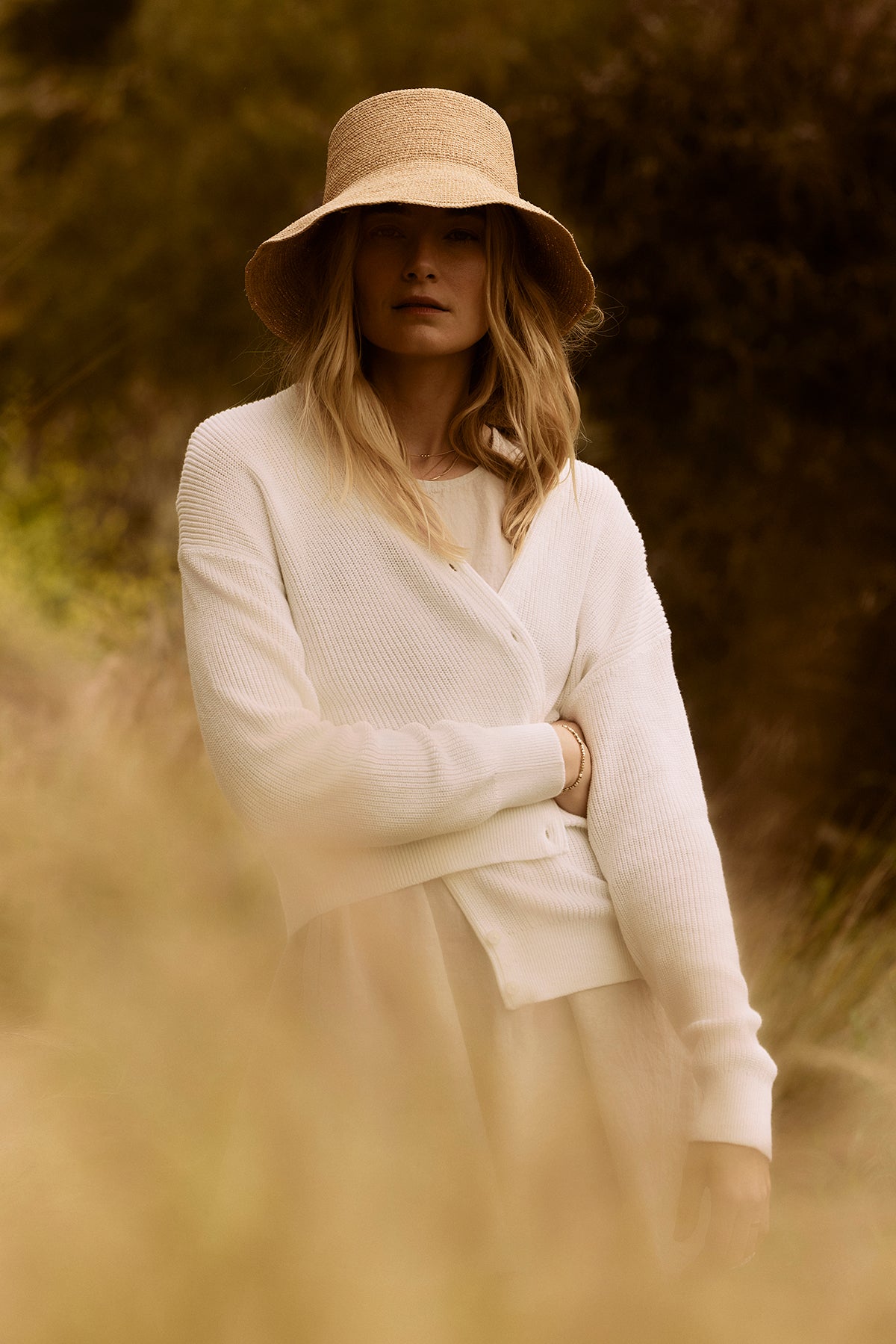   A woman in a white sweater and Velvet by Graham & Spencer CHIC CROCHET BUCKET HAT standing in a field, partially obscured by foreground foliage. 