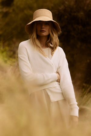 A woman in a white sweater and Velvet by Graham & Spencer CHIC CROCHET BUCKET HAT standing in a field, partially obscured by foreground foliage.