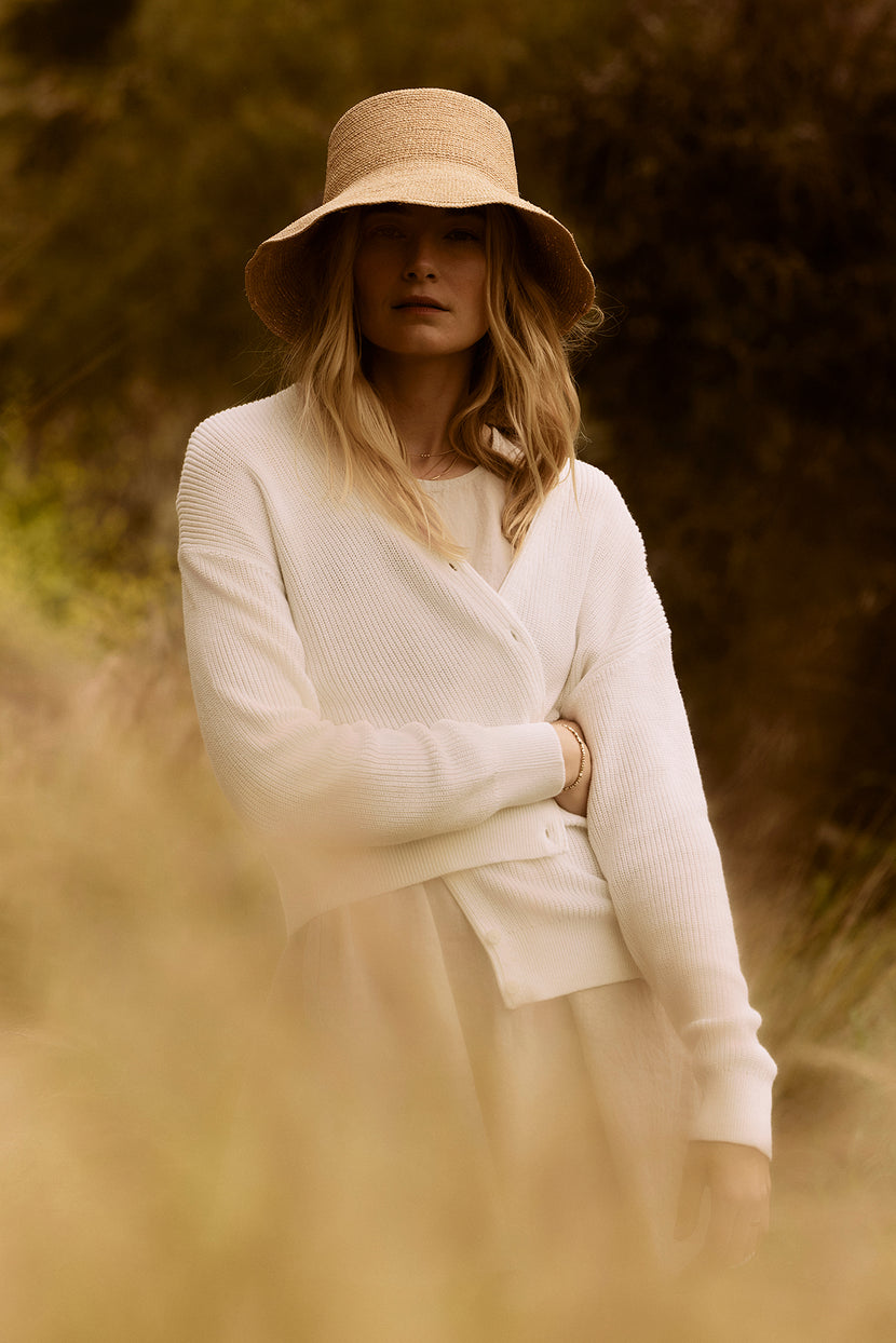 A woman in a white Velvet by Graham & Spencer TAVA cardigan and straw hat stands in a sunlit field, partially obscured by foreground foliage.