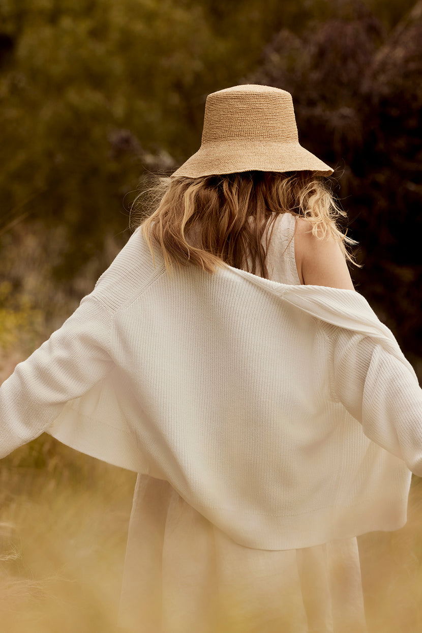 A woman with curly hair, seen from behind, wears a straw hat and a white flowing blouse in a sunlit, grassy field. She has draped over her shoulders a Velvet by Graham & Spencer Tava Cardigan.