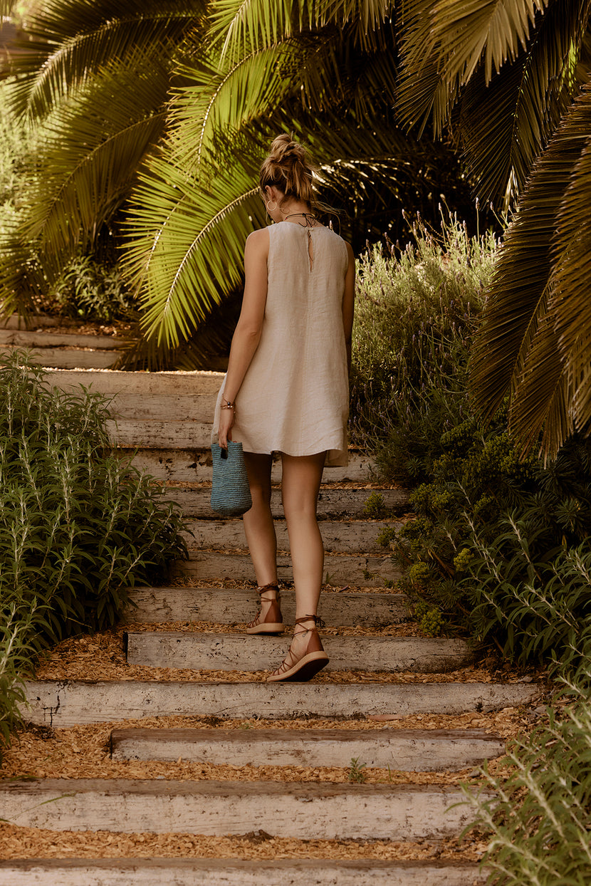 A woman in a Velvet by Graham & Spencer Tiffany Linen Dress walking up a stone staircase, surrounded by lush greenery, holding a blue bag.