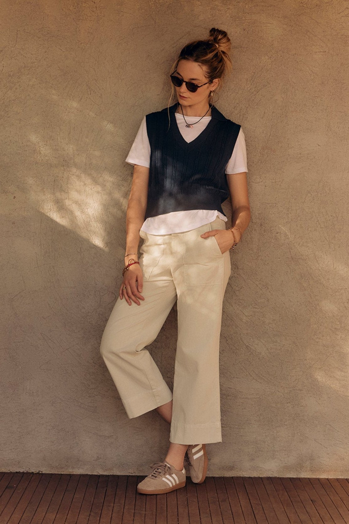 A stylish woman in sunglasses, a black vest, white shirt, and Velvet by Graham & Spencer MYA COTTON CANVAS PANT leans against a beige wall, hands in pockets.-37628822356161
