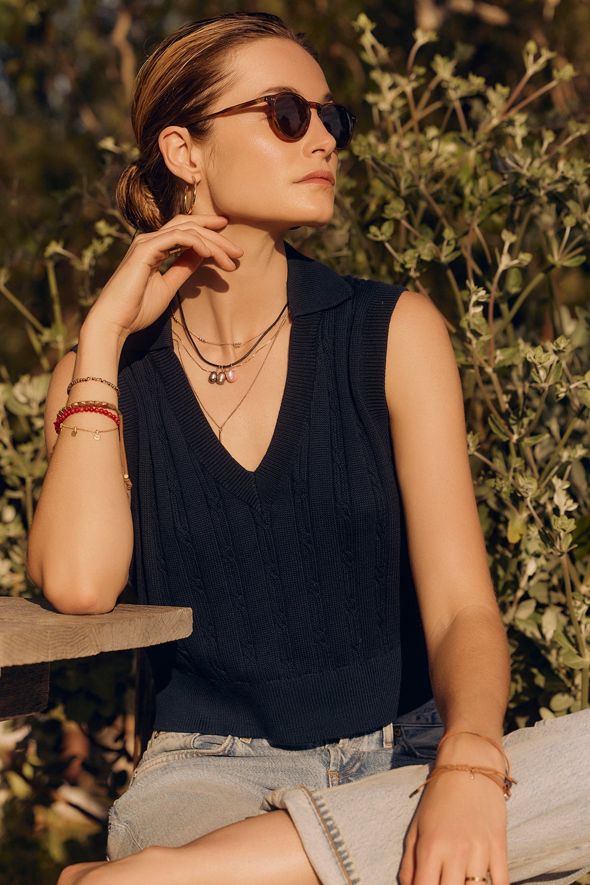   A woman wearing Velvet by Graham & Spencer sunglasses and a Wendy sweater vest sits outdoors, her hand on her neck, surrounded by green foliage. 