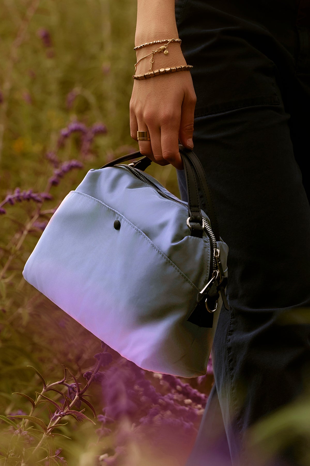   In a field of purple flowers, a person highlights the NYLON CROSSBODY BAG by Velvet by Graham & Spencer, featuring black handles and ideal for everyday use. 