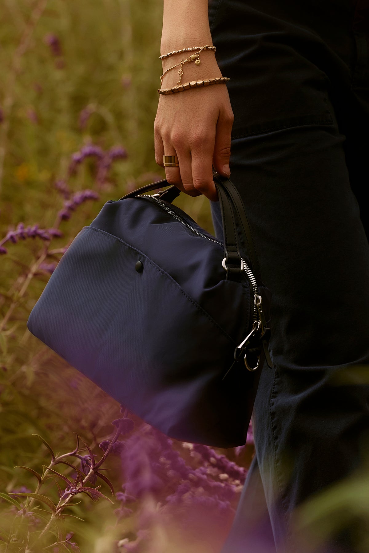   A person holds a Velvet by Graham & Spencer NYLON CROSSBODY BAG with a zipper closure by its black handle, wearing a bracelet and ring, while standing in a field of purple flowers. 