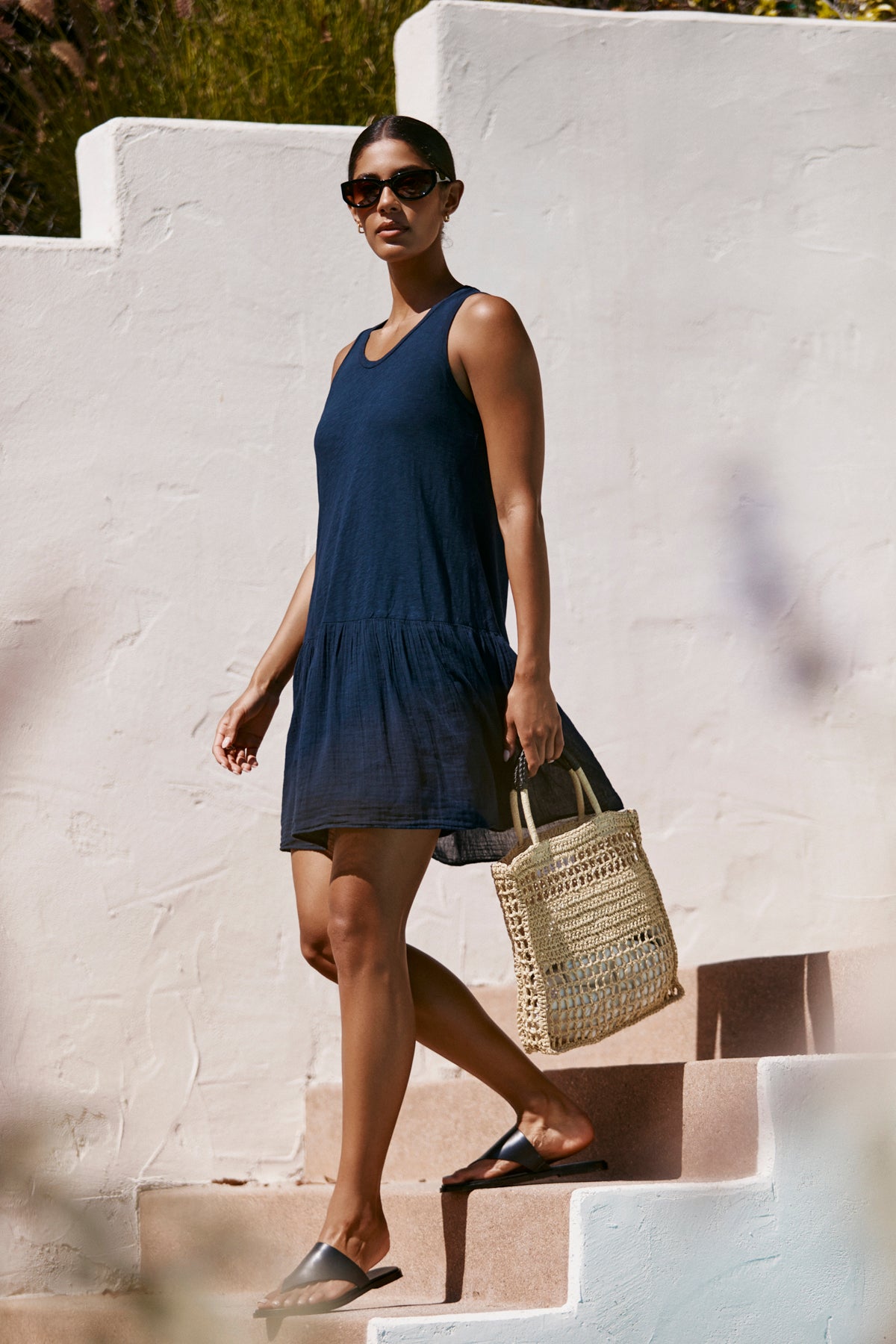 A person wearing the DENNA DRESS by Velvet by Graham & Spencer, paired with black sandals and sunglasses, walks down outdoor steps while carrying a woven tote bag.-39265825587393
