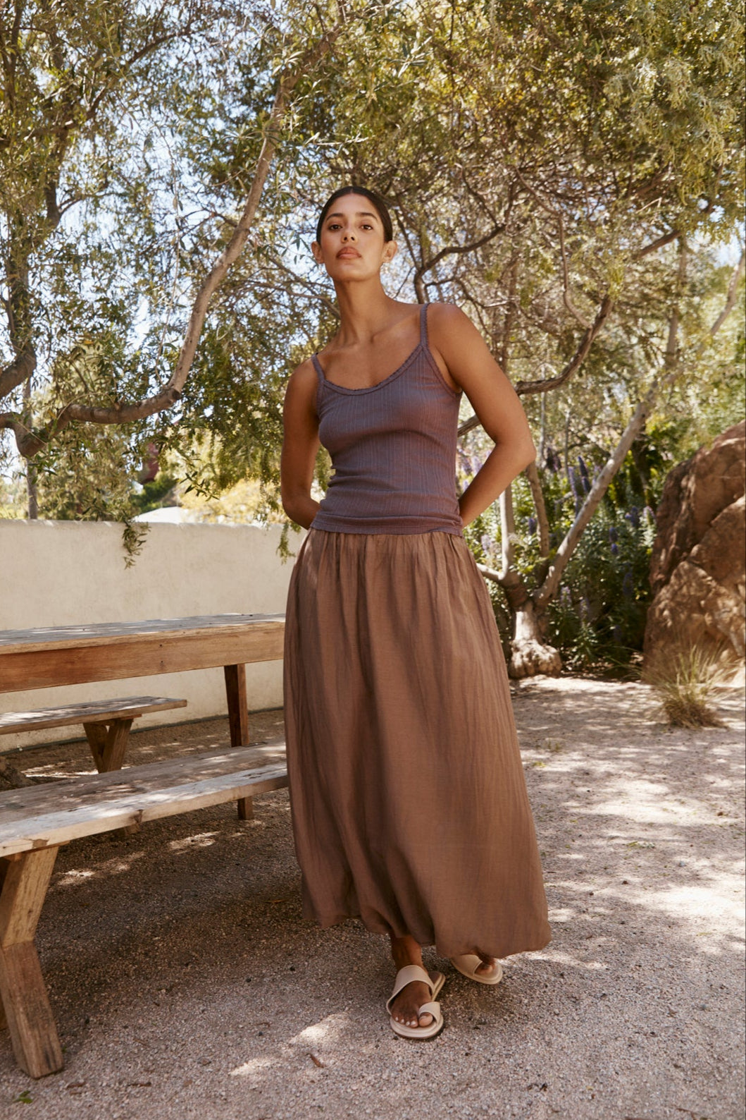 Outdoors near a wooden picnic table, a woman wears the INIKU POINTELLE TANK TOP by Velvet by Graham & Spencer with a long brown skirt and sandals, surrounded by trees and dappled sunlight.-39265837121729