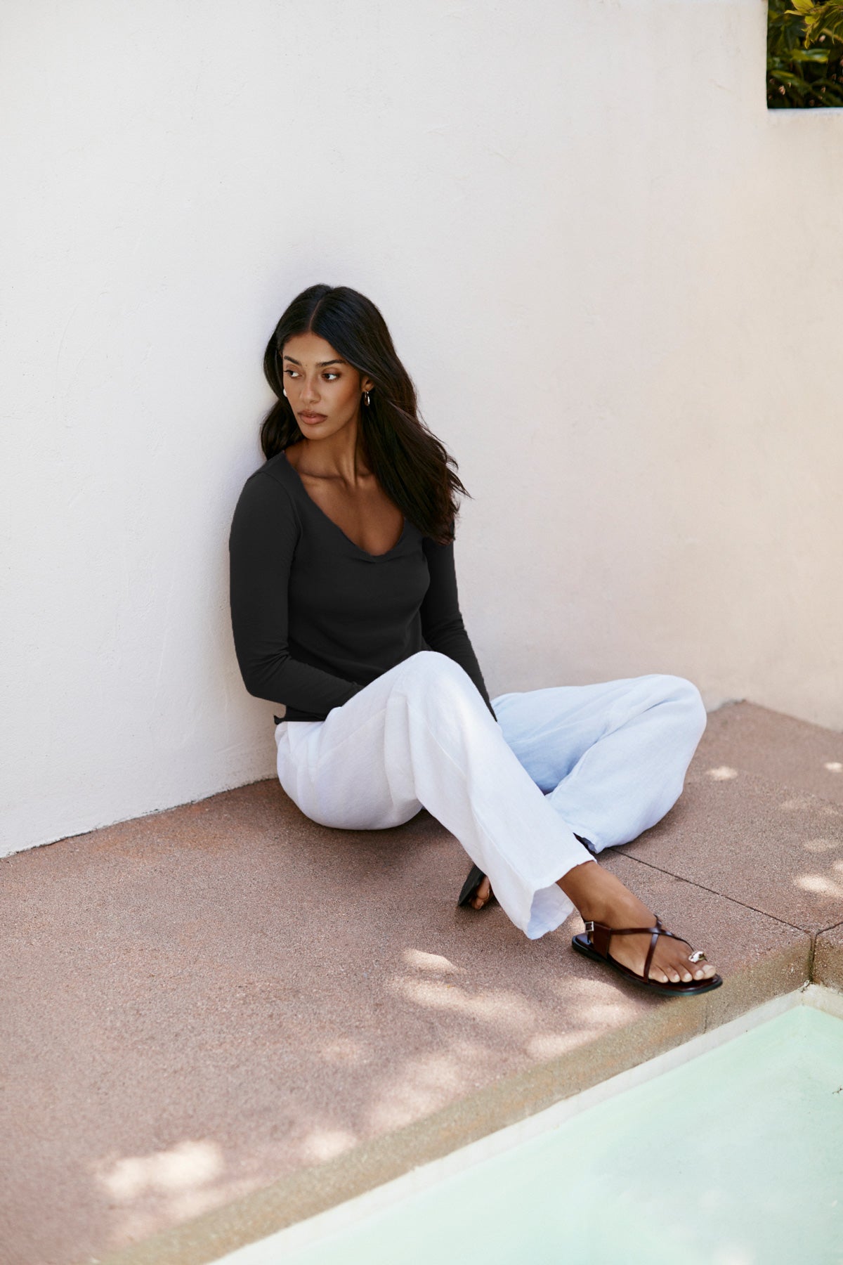 A woman wears the NOURA TEE by Velvet by Graham & Spencer—black, long sleeve, with a scoop neckline—and white pants as she sits by a pool on stone beside a white wall, gazing off to the side.-40010678206657
