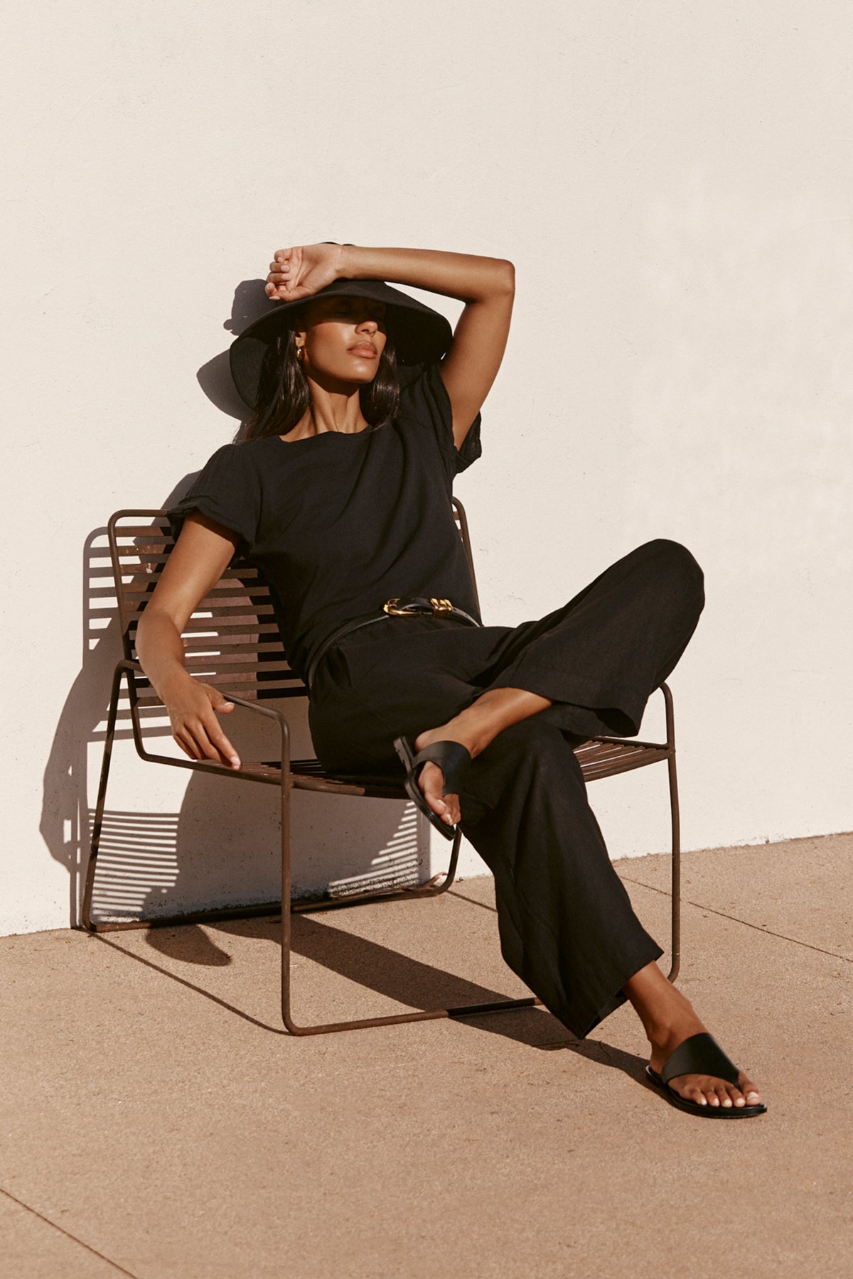   A woman in a black SOPHIA PANT by Velvet by Graham & Spencer, featuring a cropped balloon shape and elastic waist, sits outdoors in a wide-brim hat, shielding her eyes from the sun. This lightweight look is ideal for sunny days. 