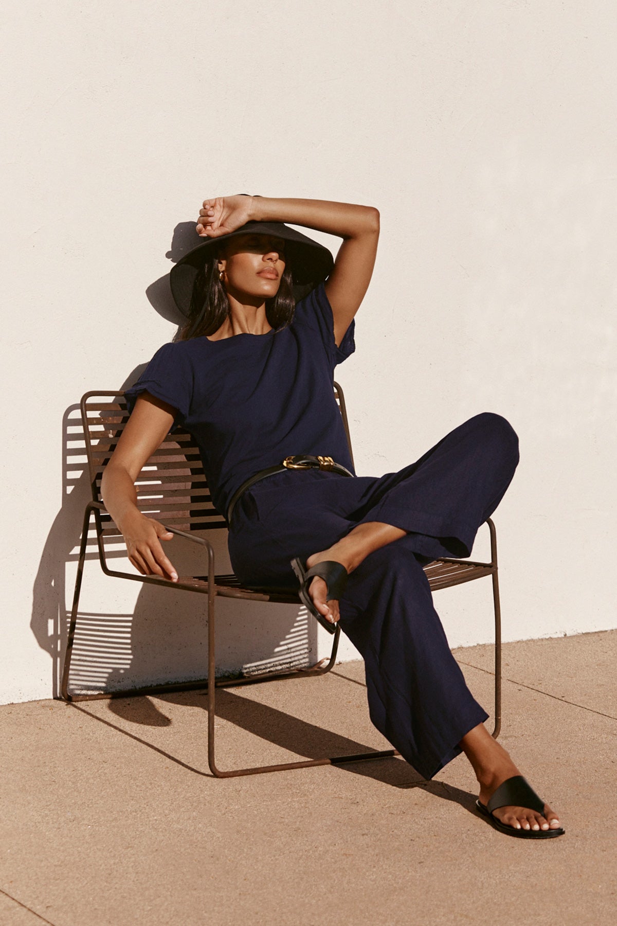   Wearing the Velvet by Graham & Spencer LOLA LINEN PANT, a person in a navy outfit and black wide-brim hat sits outdoors on a metal chair, resting one arm while shielding their eyes from the sun. 