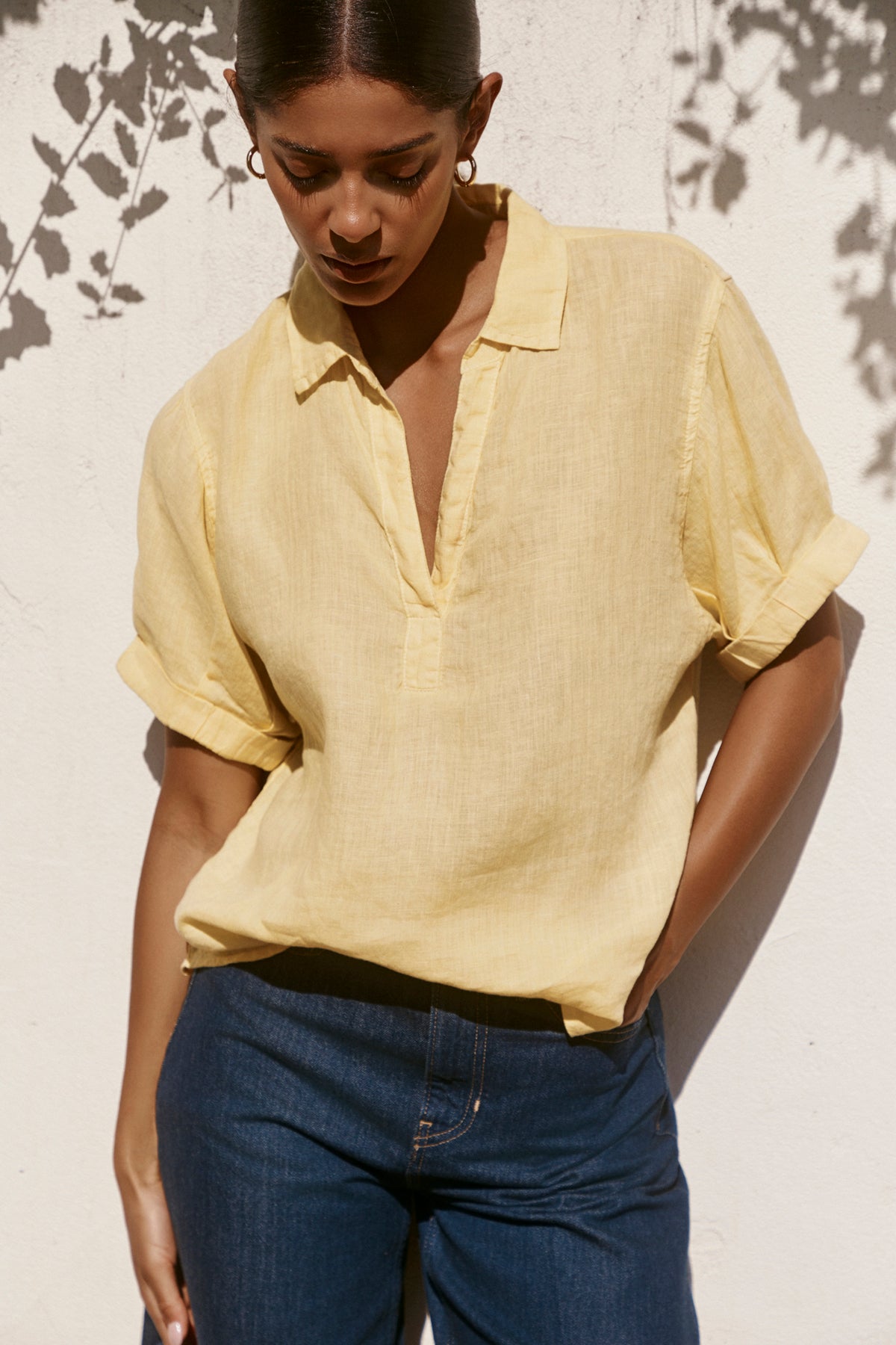   A woman wears the VIVICA LINEN TOP by Velvet by Graham & Spencer, a light yellow woven linen blouse with a V-neckline, styled with blue jeans against a sunlit white wall—ideal for warm-weather dressing. 