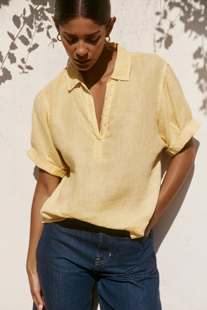 A woman wears the VIVICA LINEN TOP by Velvet by Graham & Spencer, a light yellow woven linen blouse with a V-neckline, styled with blue jeans against a sunlit white wall—ideal for warm-weather dressing.