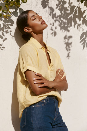 Wearing the VIVICA LINEN TOP by Velvet by Graham & Spencer with jeans, a woman stands against a sunlit wall, eyes closed and arms crossed, leaf shadows falling behind her—an effortless warm-weather style.