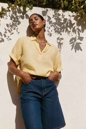 A person stands against a white wall with plant shadows, wearing the VIVICA LINEN TOP by Velvet by Graham & Spencer—a light yellow woven linen short-sleeve V-neck shirt—paired with blue denim shorts for effortless warm-weather style.
