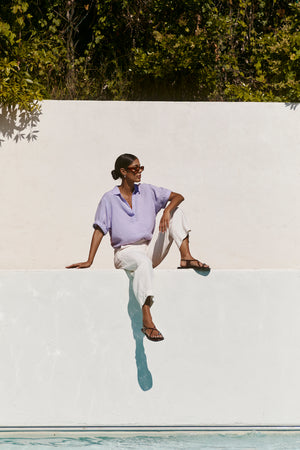 Wearing the VIVICA LINEN TOP by Velvet by Graham & Spencer, sunglasses, and white pants, a person sits on a white wall by the pool with greenery in the background—an ideal look for warm-weather dressing.