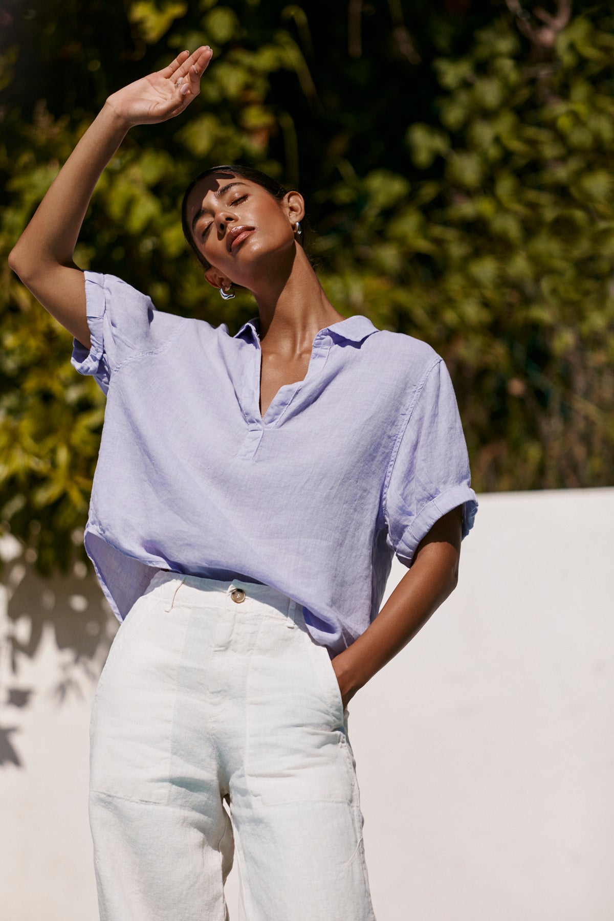 A person stands outdoors in the Velvet by Graham & Spencer VIVICA LINEN TOP, a loose lavender V-neck shirt paired with white pants. Sunlight and green foliage set a relaxed, effortless warm-weather vibe.-39266150645953
