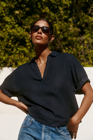 A person wears the VIVICA LINEN TOP by Velvet by Graham & Spencer—sunglasses and blue jeans complete this effortless outdoor look, with lush green foliage and a white wall in the background.