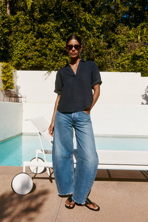 Wearing the VIVICA LINEN TOP by Velvet by Graham & Spencer, wide-leg jeans, sunglasses, and sandals, a person stands by a poolside lounge chair surrounded by greenery—a breezy look for warm-weather days.
