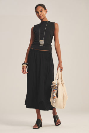 A woman wears a sleeveless black top, Velvet by Graham & Spencer's IRENE SKIRT in black cotton gauze, black heeled sandals, silver jewelry, and holds a beige bag with a patterned scarf.