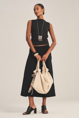 A woman stands against a plain background wearing the IRENE SKIRT by Velvet by Graham & Spencer, paired with a black sleeveless top, jewelry, black heeled sandals, and holding a large beige bag.