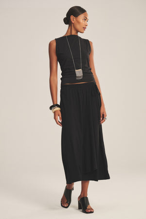 A woman stands against a plain light background, wearing the IRENE SKIRT by Velvet by Graham & Spencer—a black midi wrap skirt—paired with a sleeveless black top, black heeled mules, silver necklace, and bangles.