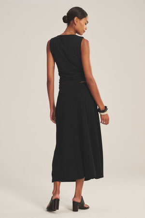 A woman with her hair tied back faces away, wearing the IRENE SKIRT by Velvet by Graham & Spencer with a black cotton gauze sleeveless top, black heeled sandals, and bracelets.